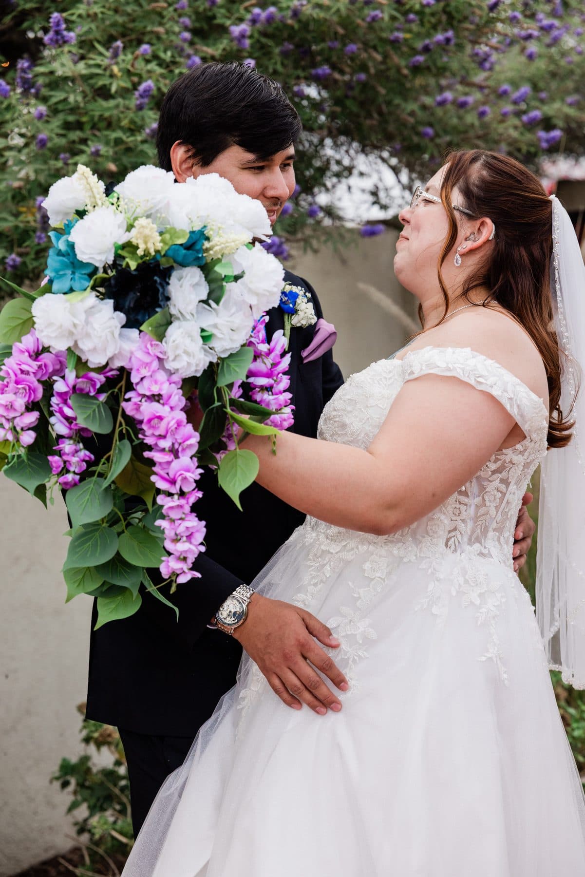 The groom looking at the bride beside her bouquet of white and lavender blooms.